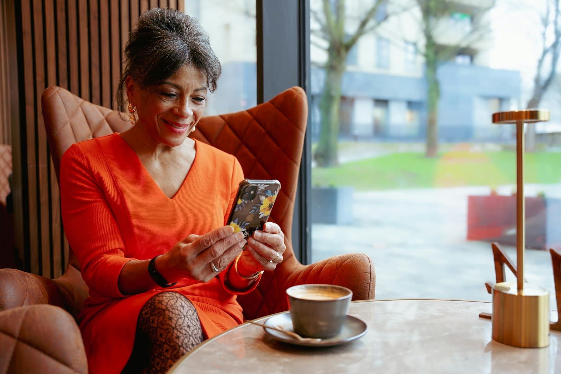 Une patiente assise dans un fauteuil près d’une baie vitrée, son téléphone en main, un café sur la table.